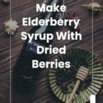 A bottle of elderberry syrup, a fluted dish with dried berries, and a honey dipper on a wooden table with scattered flowers. Text overlay: “How to Make Elderberry Syrup With Dried Berries.”.