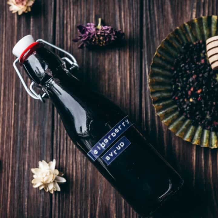 A glass bottle labeled “elderberry syrup” sits on a wooden surface beside dried flowers, a plate of elderberries, and a honey dipper—suggesting the perfect setup for creating your own homemade elderberry syrup recipe.