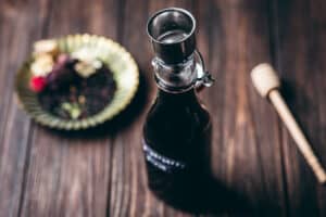 A glass bottle with a metal pour spout sits on a wooden table near a honey dipper and a plate with assorted dried herbs—perfect inspiration for trying your own elderberry syrup recipe.