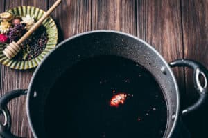 A pot filled with dark liquid, reminiscent of an elderberry syrup recipe, sits on a wooden surface, with a single floating spice. A plate with dried flowers, spices, and a honey dipper is placed nearby.