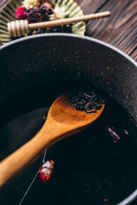 A wooden spoon stirs a dark liquid with spices in a black pot—an elderberry syrup recipe in the making; a honey dipper and herbs are blurred in the background.