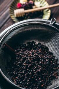 A close-up of dried elderberries in a metal strainer, with a cinnamon stick visible and a blurred background showing a plate and honey dipper—perfect ingredients for your favorite elderberry syrup recipe.
