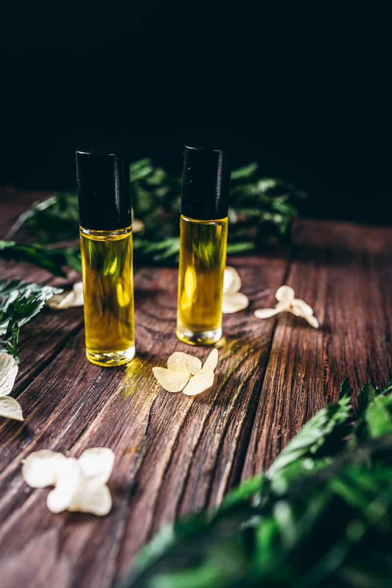 Two small roll on bottles filled with yellow oil resting on dark wooden table topped with dried plants.