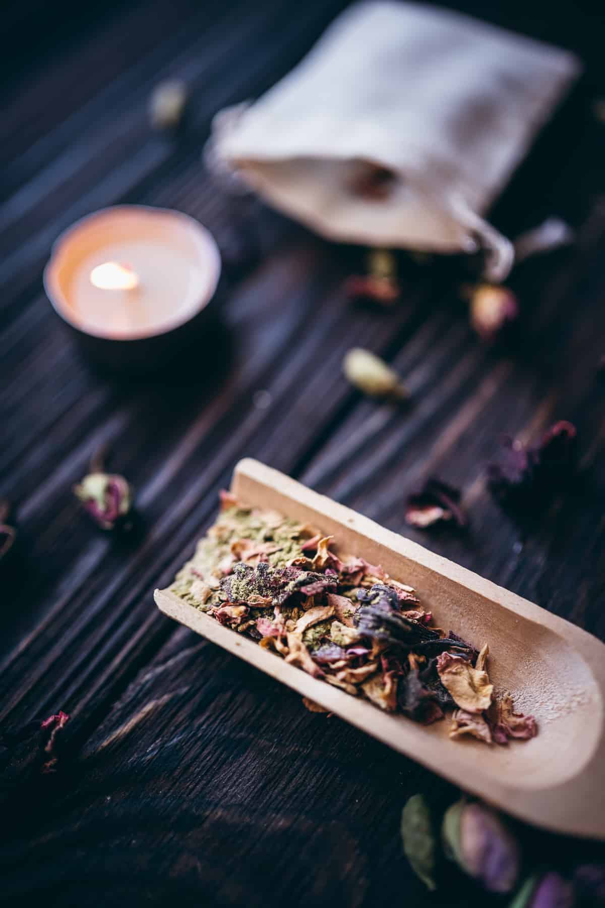 A wooden spoon filled with dried herbs and flowers resting on a dark table next to a lit candle.