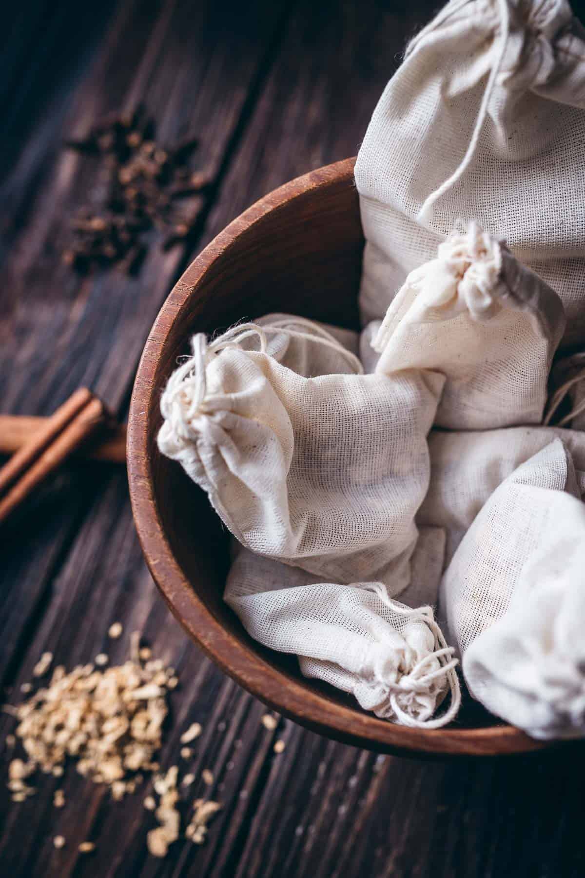 A wooden bowl filled with closed white muslin bags rests on a dark table top.