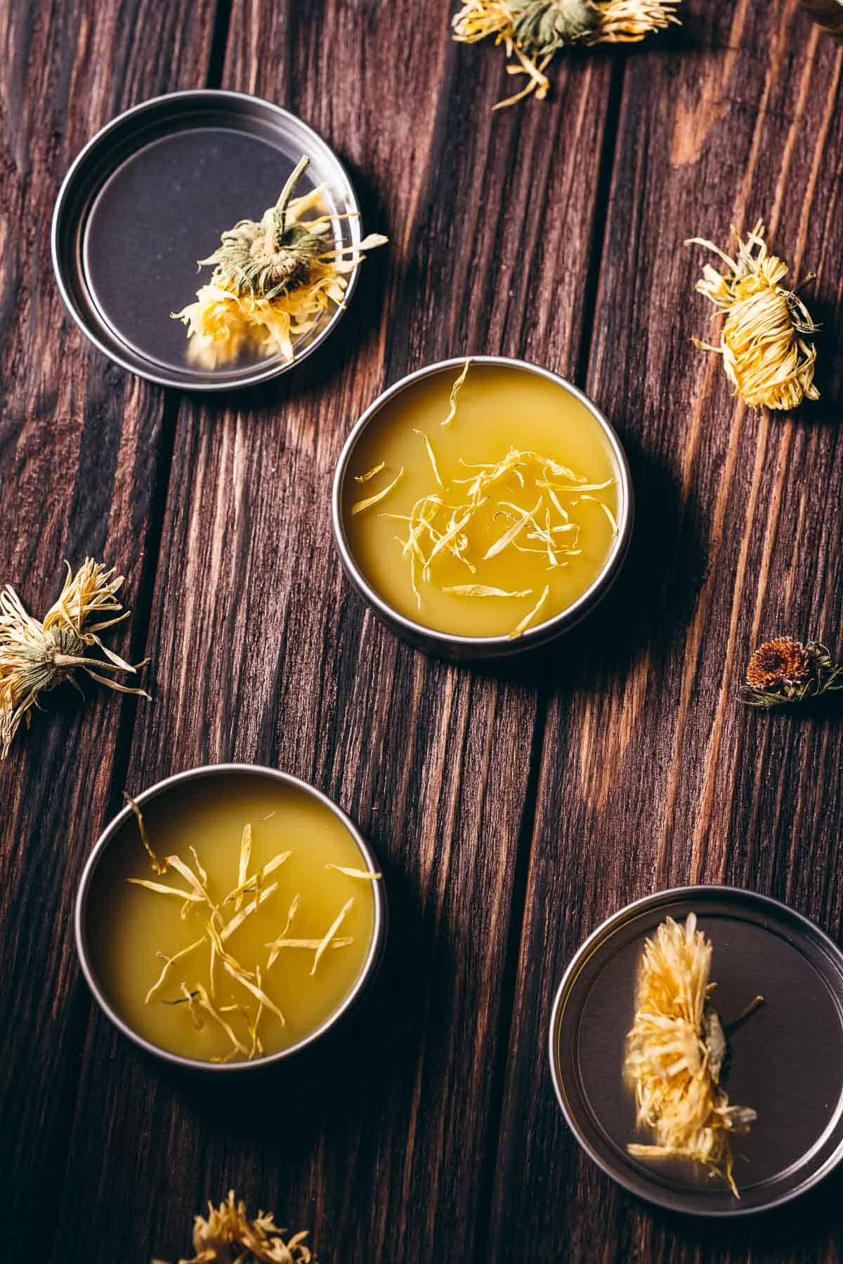 Top view of open metal salve tins filled with a yellow balm topped with golden flower petals.