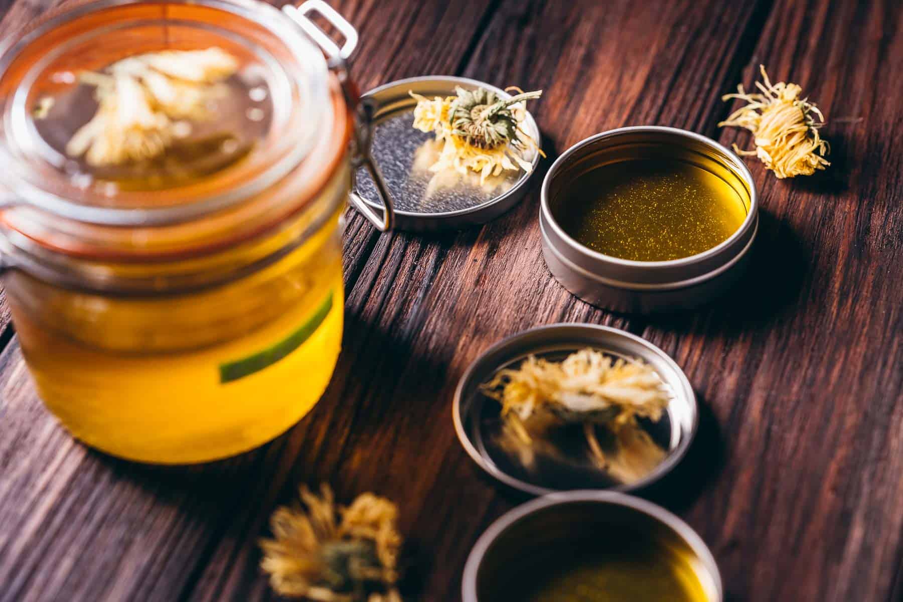 A clear glass jar filled with oil and a green label that says calendula, rests next to open metal tins filled with golden liquid.