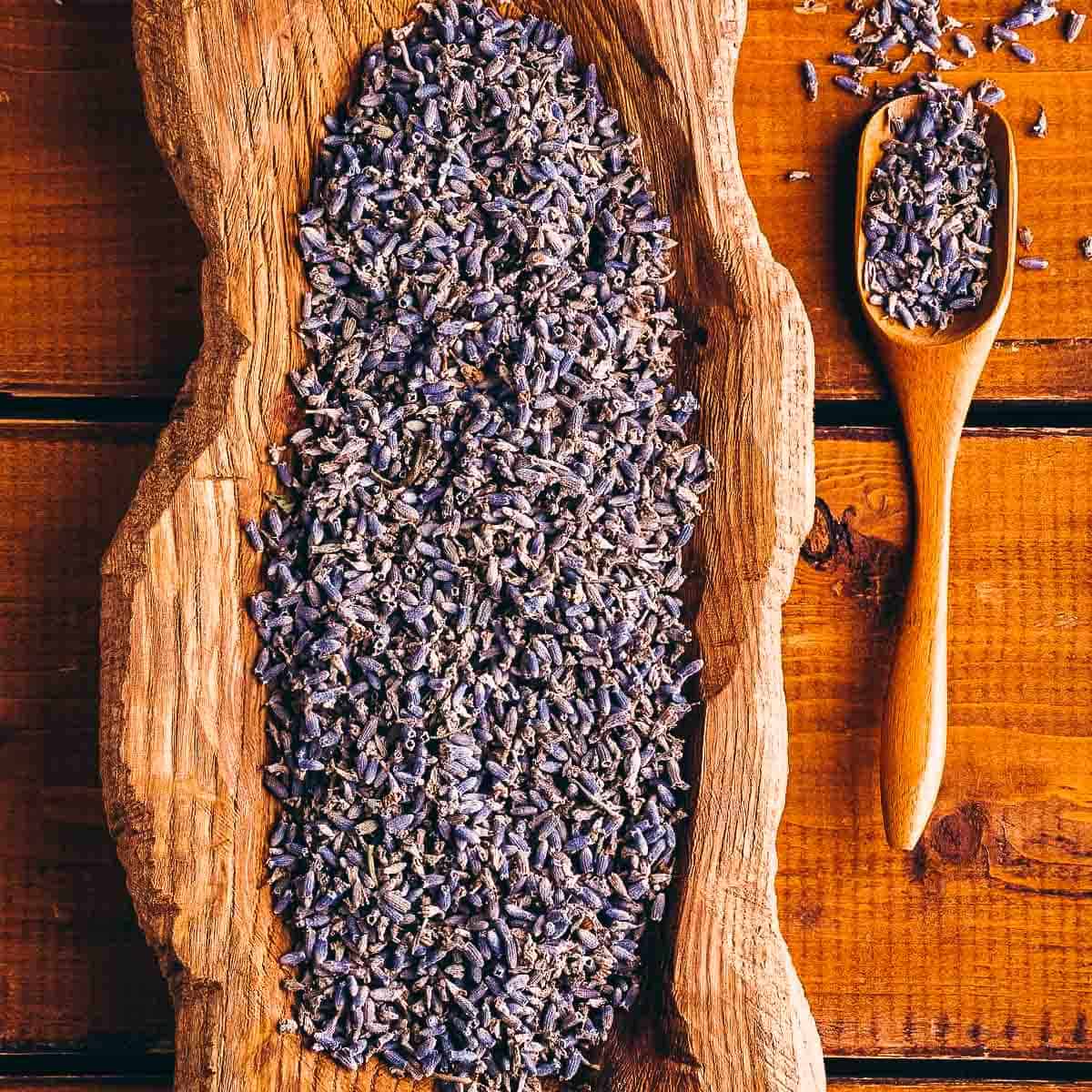 A wooden bowl filled with dried lavender buds rests on a wooden table next to a wooden spoon.