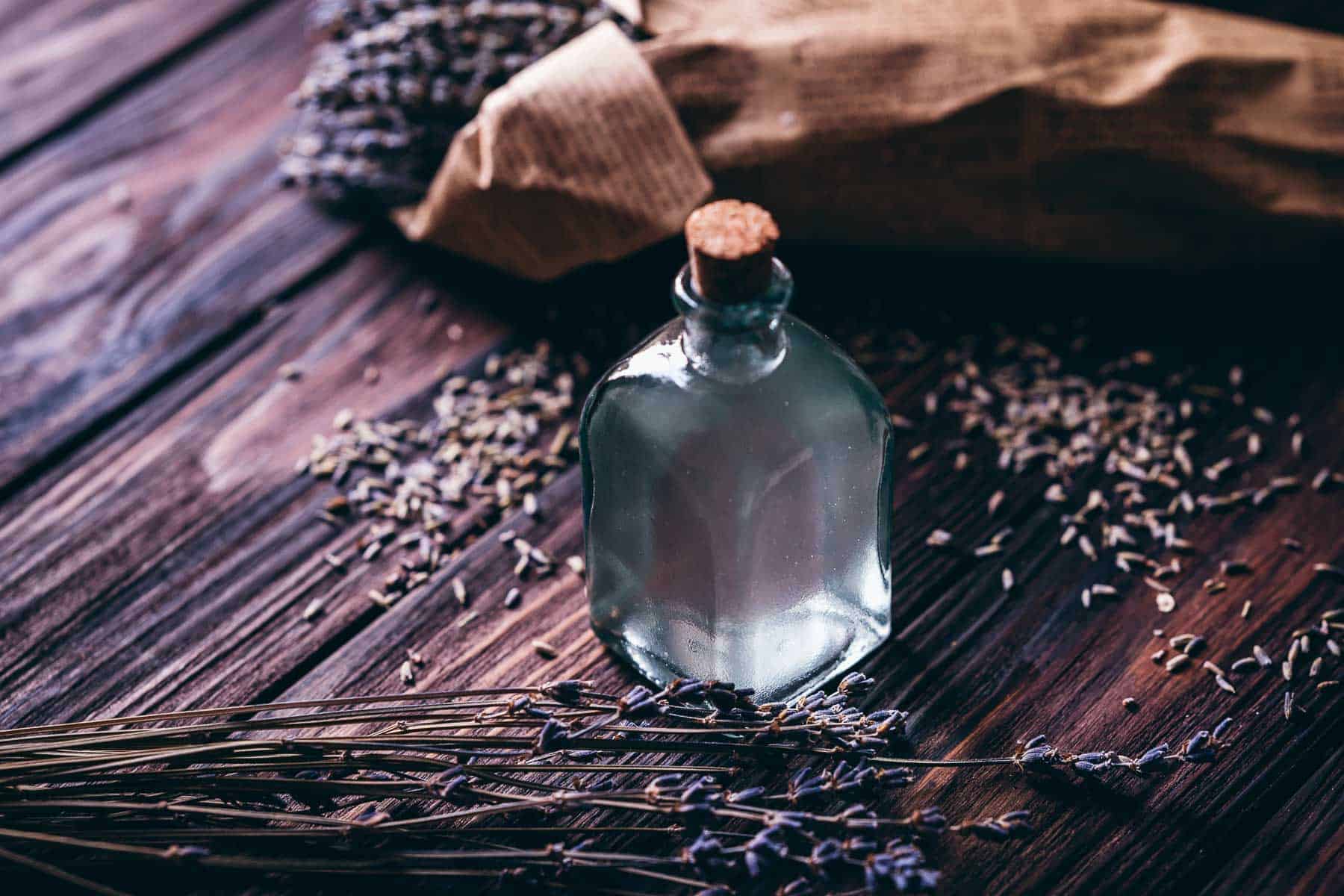 A clear glass bottle of lavender water with a cork top rests on a wood table next to dried lavender buds.