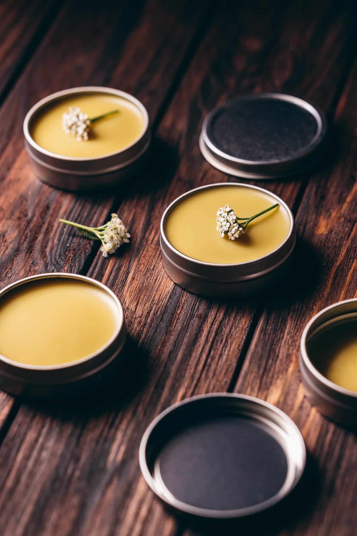 Close view of a silver salve tin fillwed with a yellow balm and topped with a small white yarrow flower.
