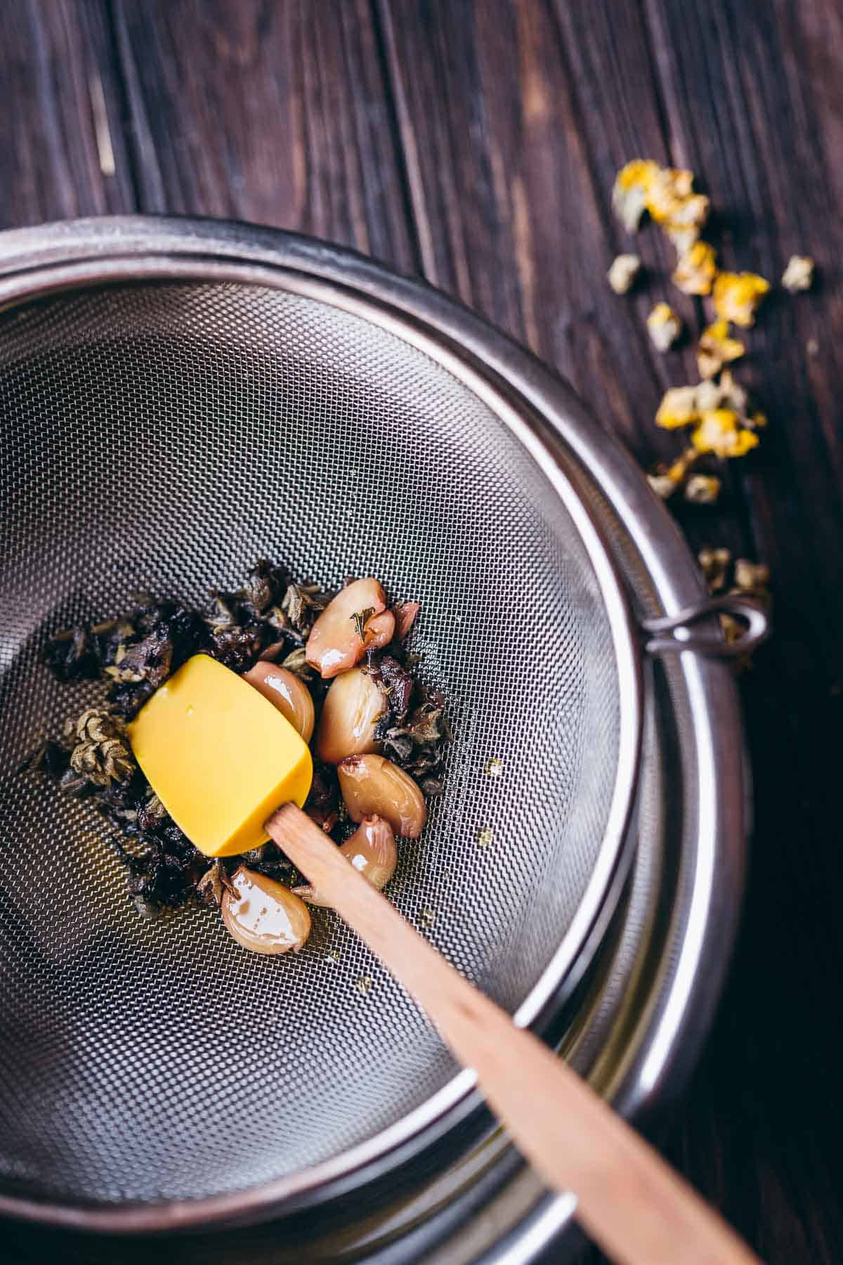 A metal mesh strainer filled with garlic cloves and mullein flowers rests on a big silver mixing bowl.