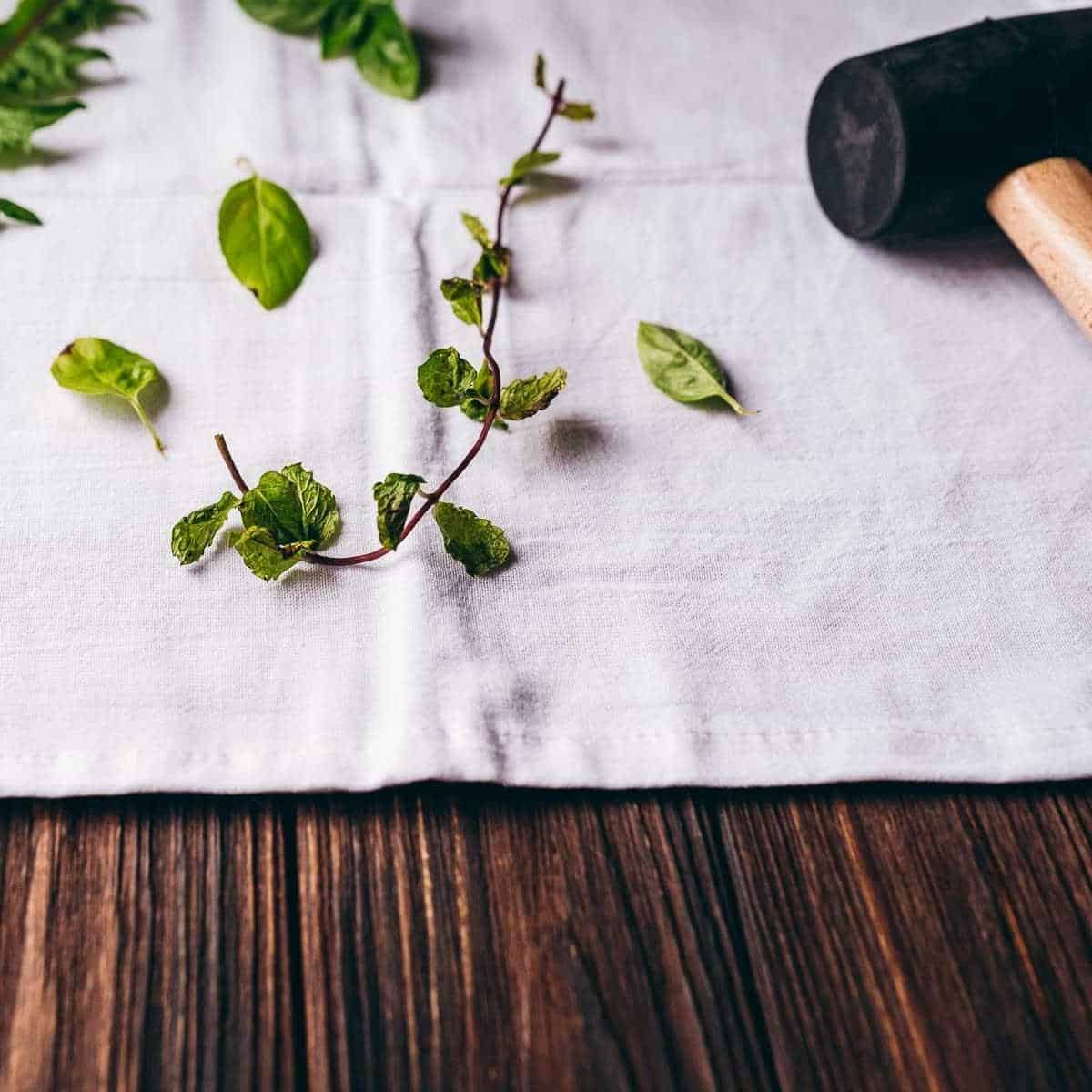 White fabric rests on a dark table next to fresh green leaves and a black rubber mallet.