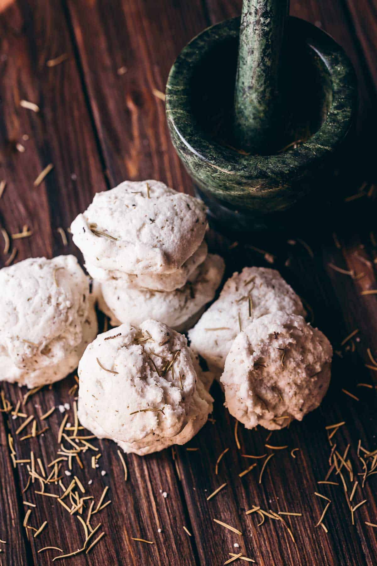 A pile of homemade rosemary bath truffles resting on a dark wooden table next to a dark green mortar and pestle.