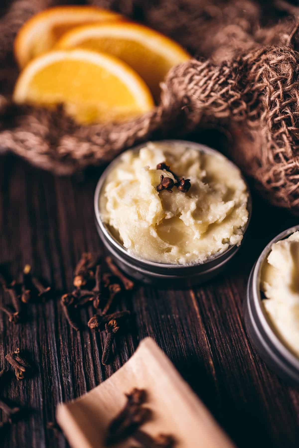 Close shot of a metal tin of whipped body butter topped with whole cloves.