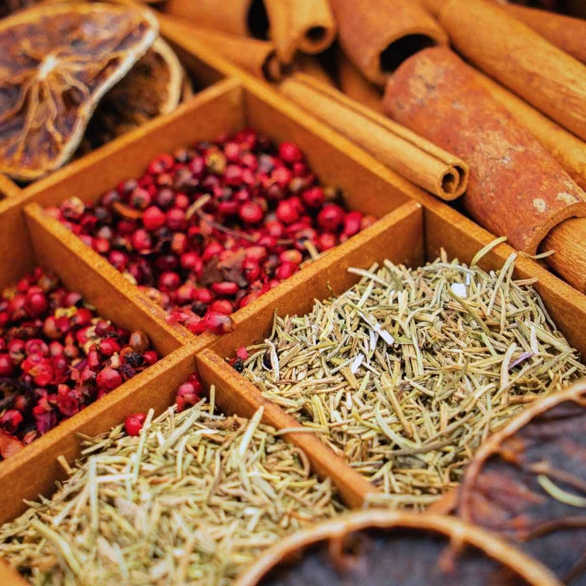 A wooden box contains dried herbs and spices, including pink peppercorns, rosemary, and cinnamon sticks—each known as herbs for prosperity—neatly separated into sections.