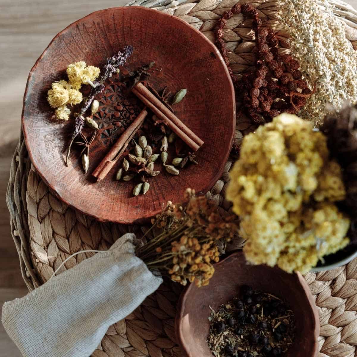 A wooden bowl holds cinnamon sticks, cardamom, and dried flowers—traditional herbs for prosperity—resting on a woven mat beside a linen pouch, yellow flowers, and another bowl filled with dried herbs.