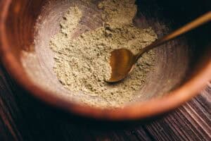 A metal spoon rests in a wooden bowl containing a coarse, green-brown powder used to make incense cones.