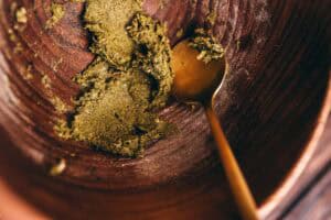 A brass spoon rests in a wooden bowl with a clump of crushed green herbs for making incense cones.