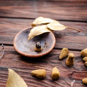A burning incense cone in a small ceramic bowl, surrounded by several unlit incense cones for those learning how to make incense cones, with dried leaves on a wooden surface.