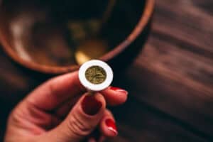 A hand holds a small, white smoking pipe bowl filled with dried herbs used to make incense cones, with a wooden bowl in the background.