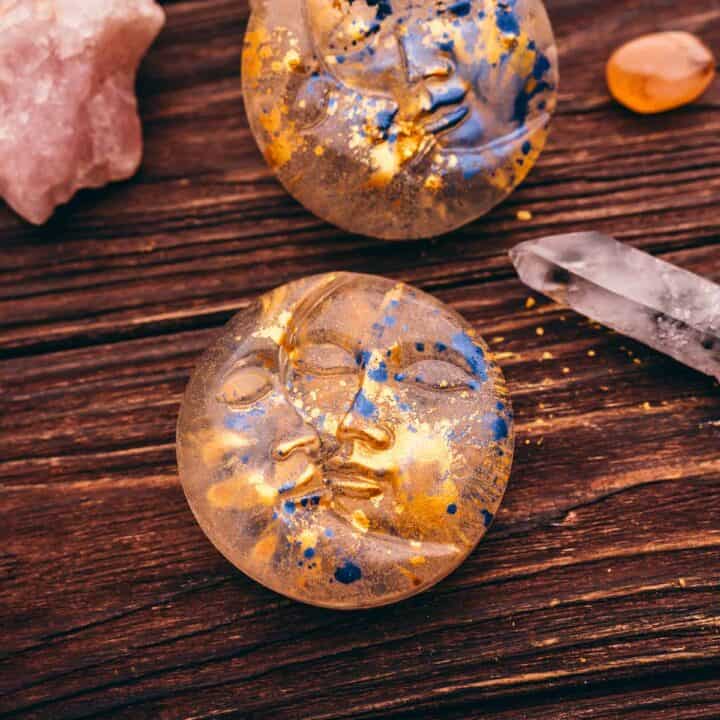 Two round face sculptures with blue and gold flecks, a quartz crystal, and other minerals rest on a wooden surface, evoking the charm of a celestial soap recipe.