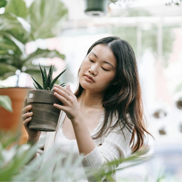 A woman in a white sweater examines a potted plant in a lush, green plant store.