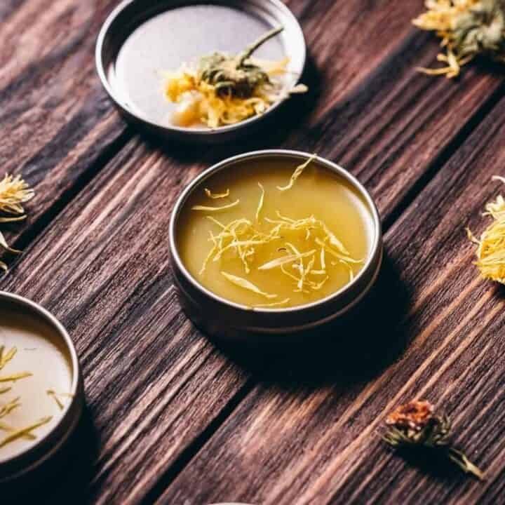 Tin containers with herbal balm on a wooden surface, surrounded by scattered dried yellow flowers.