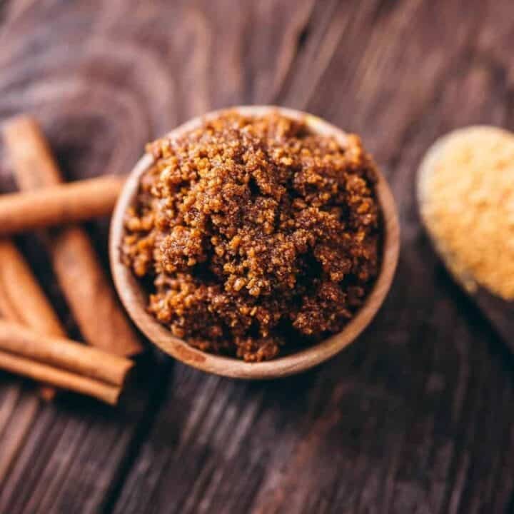 A bowl of brown sugar granules next to cinnamon sticks and a wooden spoon with raw sugar on a wooden surface.