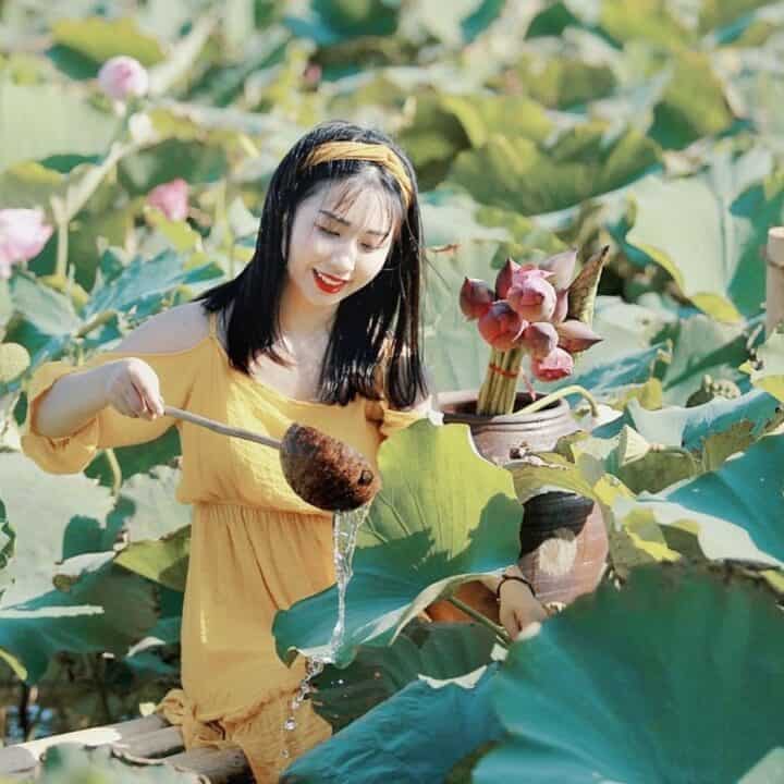 Woman in a yellow dress waters plants with a ladle in a lush field of large green leaves and pink flowers.
