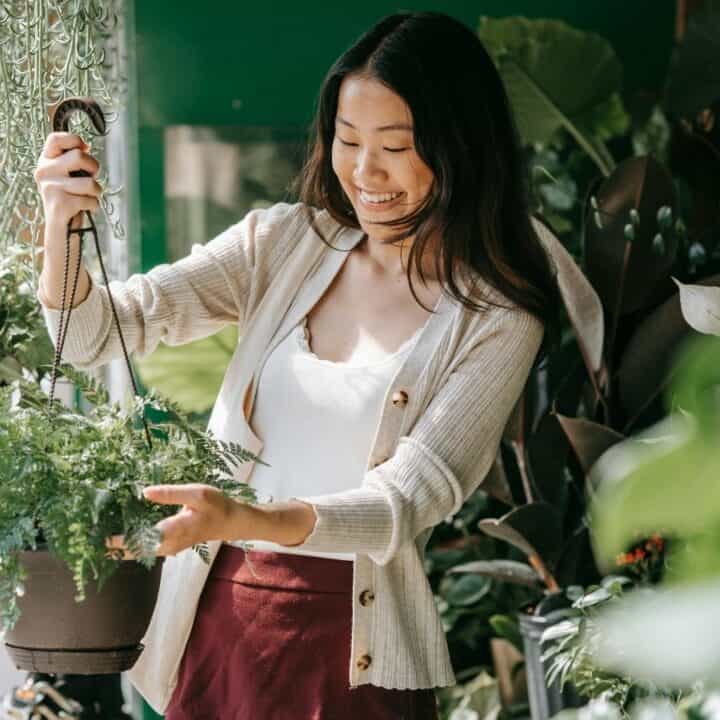 A woman in a cardigan holds a hanging potted plant while smiling, surrounded by lush greenery.