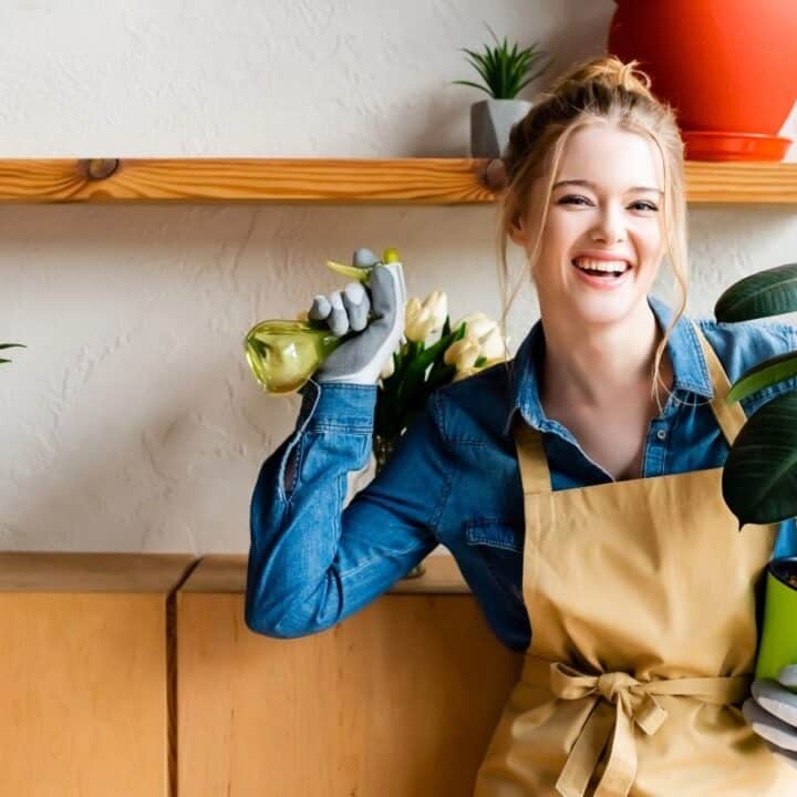 A person in a denim shirt and apron holds a watering can and a potted plant, smiling in a room with various plants on shelves.