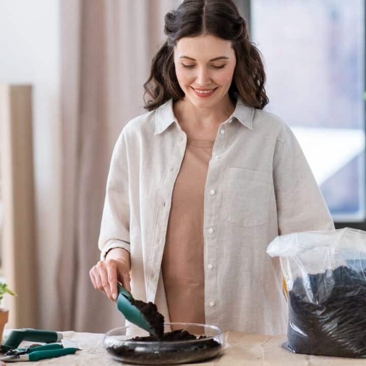 A woman smiles while scooping soil from a bag into a glass container on a table, surrounded by potted plants in a well-lit room.