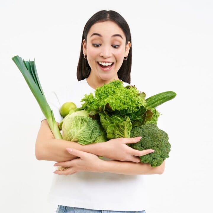 A woman in a white shirt holds a bundle of fresh vegetables, including lettuce, broccoli, and leeks, against a plain white background.
