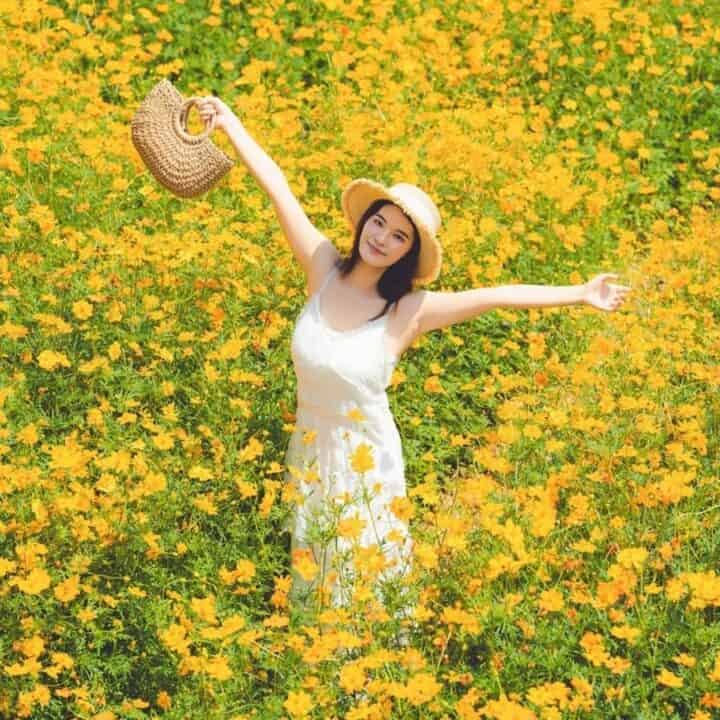 A woman in a white dress and hat stands with arms outstretched in a field of yellow flowers, smiling and holding a straw bag.