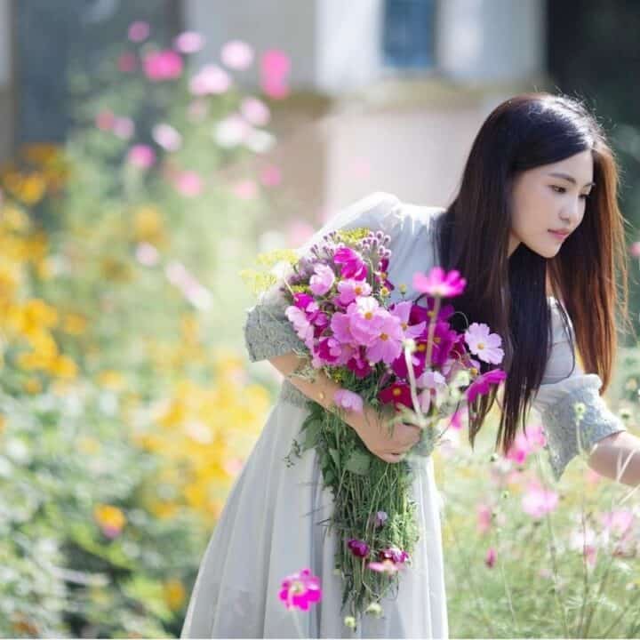 A woman in a light dress holds a bouquet of flowers while leaning forward to touch pink blossoms in a colorful garden.
