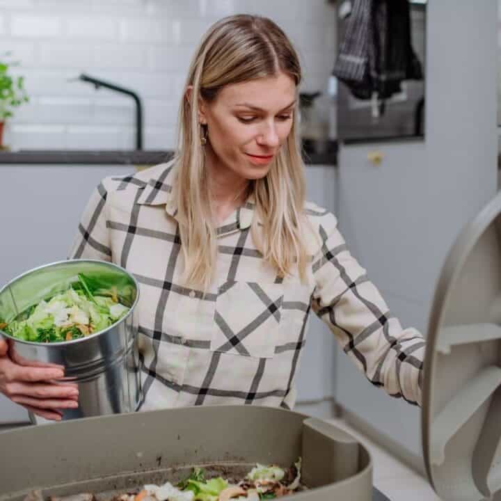 A woman in a kitchen disposes of food scraps into a compost bin. She holds a metal container with vegetable waste.