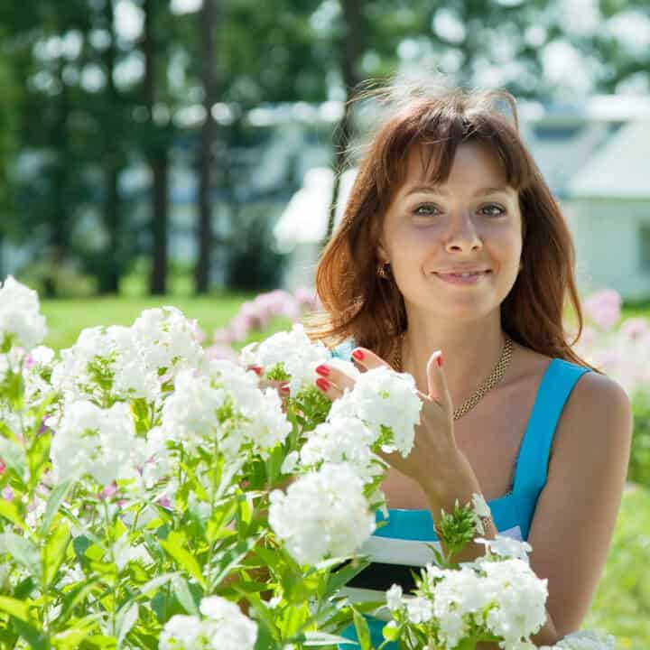 A woman in a blue dress stands outdoors among white flowers, smiling at the camera with greenery and buildings in the background.
