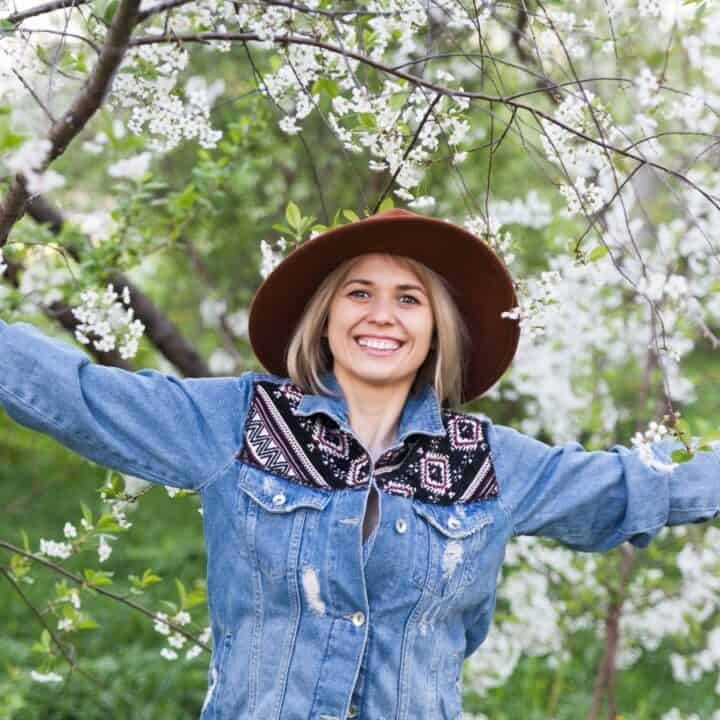 Woman in a denim jacket and brown hat stands outdoors with arms outstretched, smiling in front of blooming trees with white flowers.