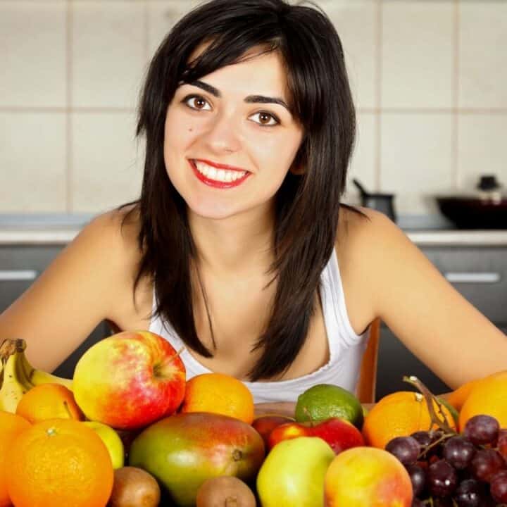 A woman with dark hair and a white top sits at a table in a kitchen, smiling behind a large assortment of fresh fruits including pineapple, oranges, apples, bananas, and grapes.