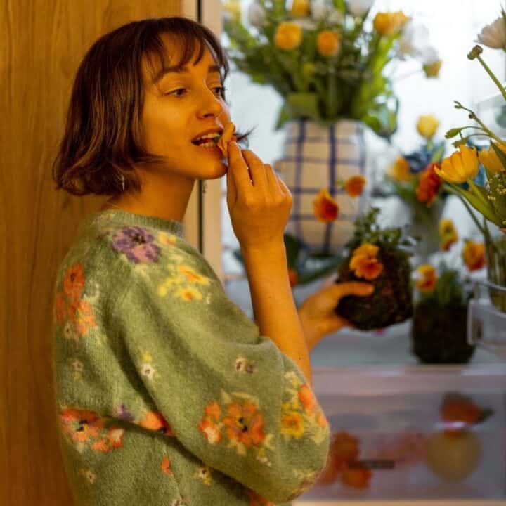 A woman applies lipstick while standing in front of an open refrigerator filled with flowers and vases.