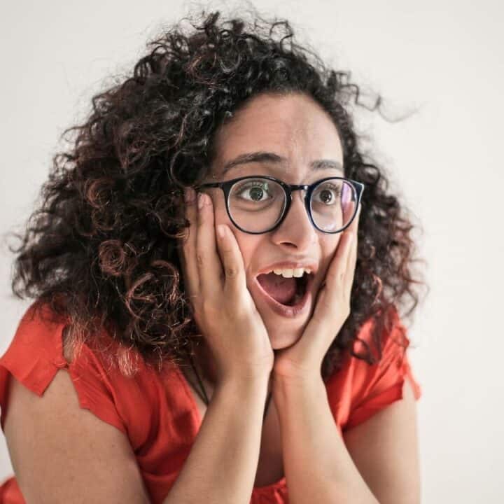 A woman with curly hair and glasses, wearing a red shirt, holds her face with both hands and looks surprised or excited against a plain background.
