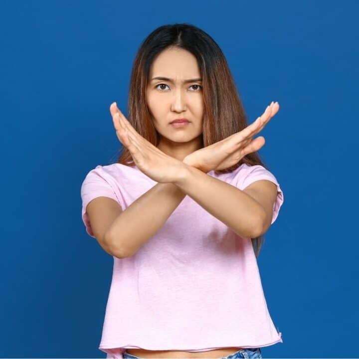 Woman standing in front of a blue background making an "X" with her arms in front of her chest, indicating refusal or prohibition.