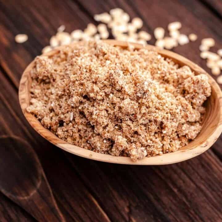 A wooden bowl filled with brown, grainy sugar sits on a dark wooden surface. Scattered sugar granules are visible around the bowl.
