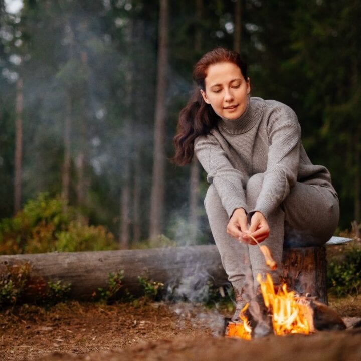 A person in gray clothing crouches near a campfire in a forest, roasting a marshmallow on a stick. Large trees are visible in the background.