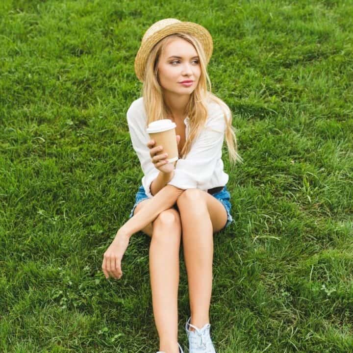 Woman sitting on grass, wearing a straw hat and holding a coffee cup. She has long blonde hair, a white shirt, and denim shorts.
