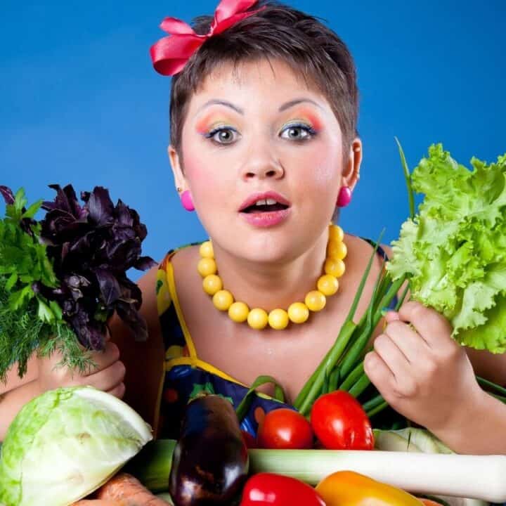 A surprised person holding leafy vegetables with various colorful vegetables spread on a table in front. Blue background.