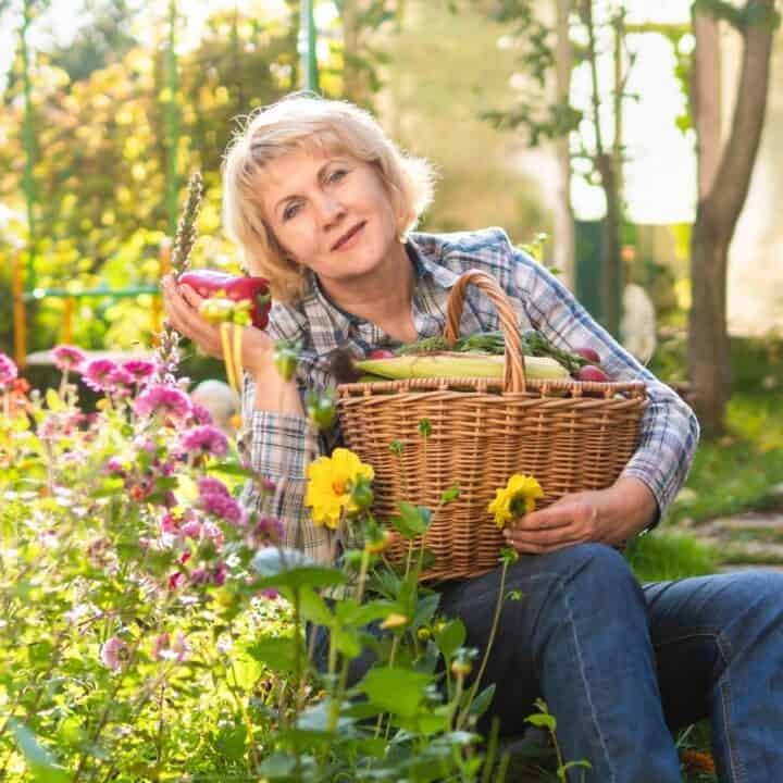A woman sits in a garden holding a large basket of vegetables and flowers, surrounded by blooming plants and greenery.