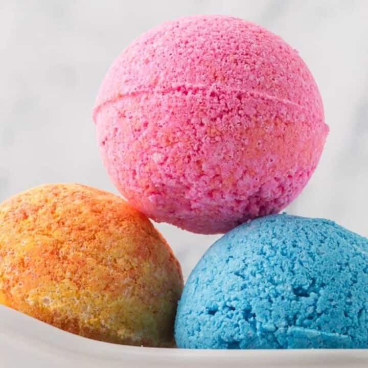 Three colorful bath bombs—pink, yellow-orange, and blue—stacked in a white dish against a light, blurred background.