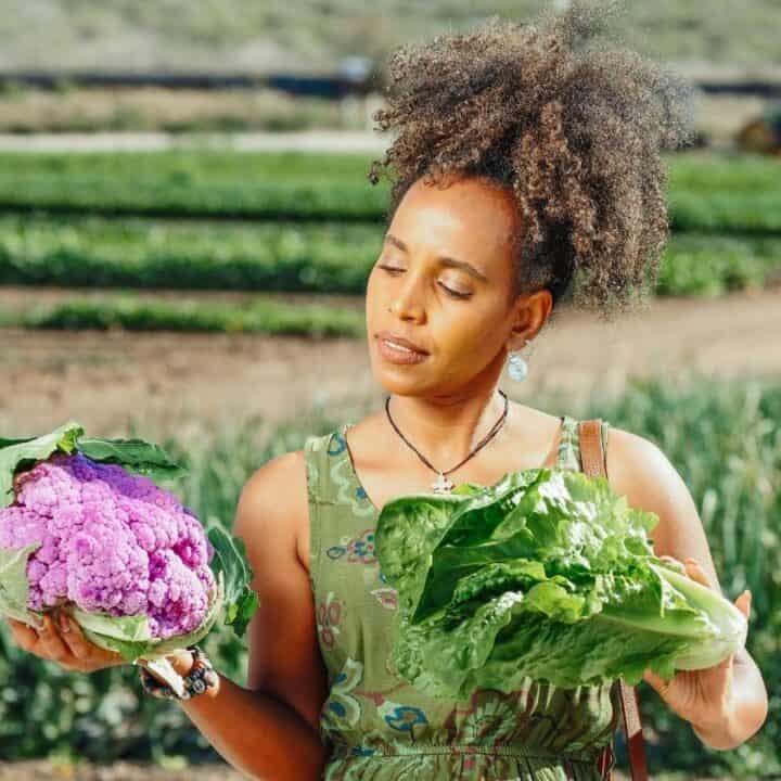 A woman stands in a field holding a purple cauliflower in one hand and green lettuce in the other, contemplating her choice.