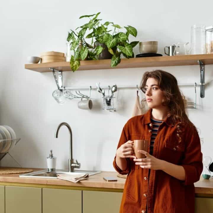 Woman in a brown shirt holds a mug, standing in a modern kitchen with plants, dishes, and utensils on shelves and countertop.
